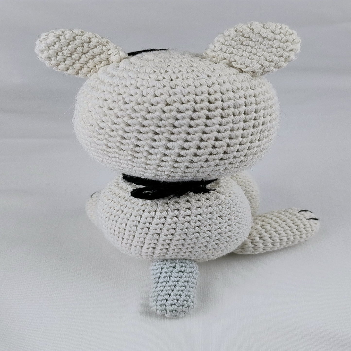 Rear view of a handmade crochet amigurumi cat sitting solo. The focus is on the rounded shape of the body, the back of the ears, and a long yarn tail resting on the ground.